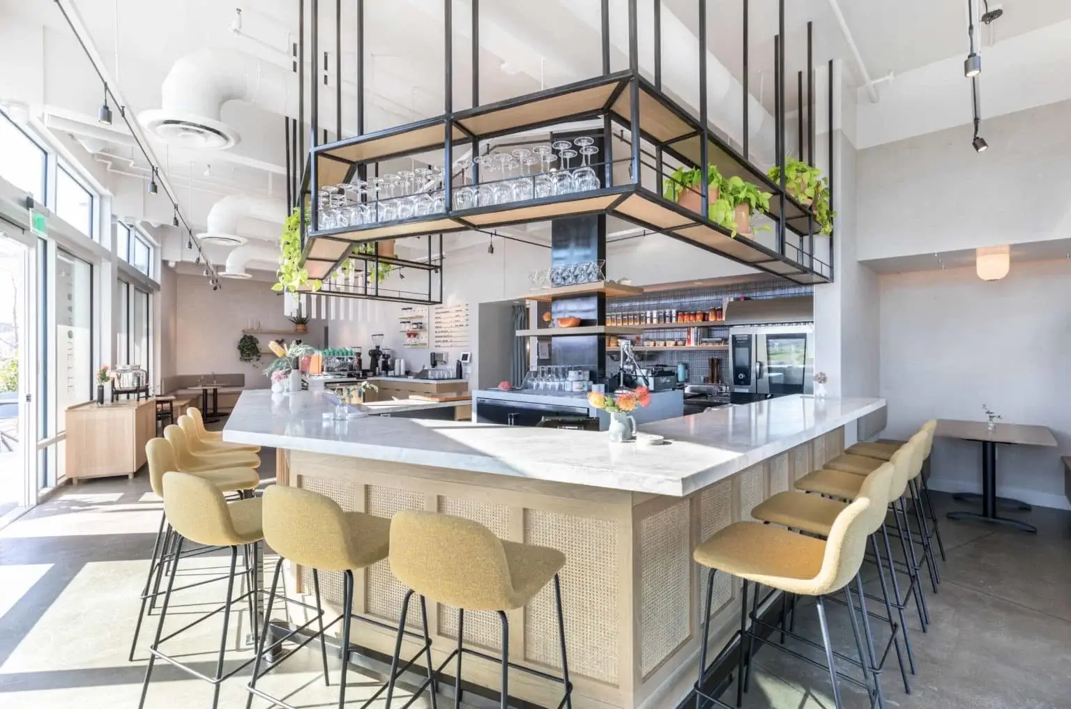 Franquette_WEB-5-1536x1017 Modern bar in The Bridge District with a white marble countertop, yellow cushioned bar stools, hanging shelves displaying glasses and plants, and bright natural lighting from large windows.