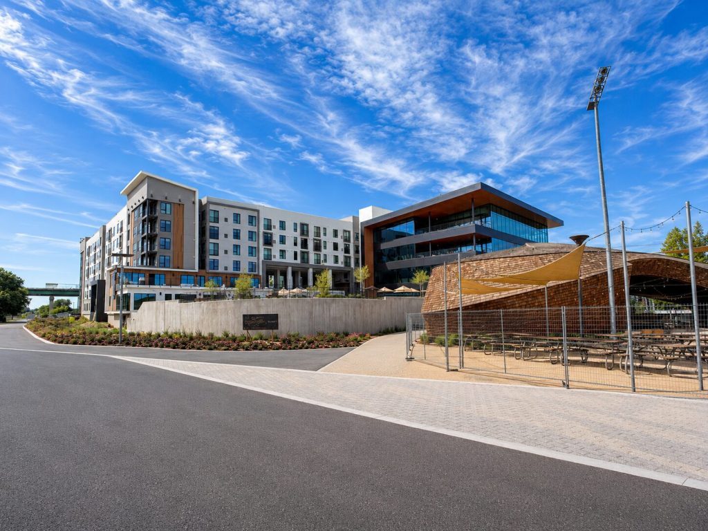 Modern multi-story building complex with adjacent open-air wooden pavilion and landscaped grounds under a partly cloudy blue sky.
